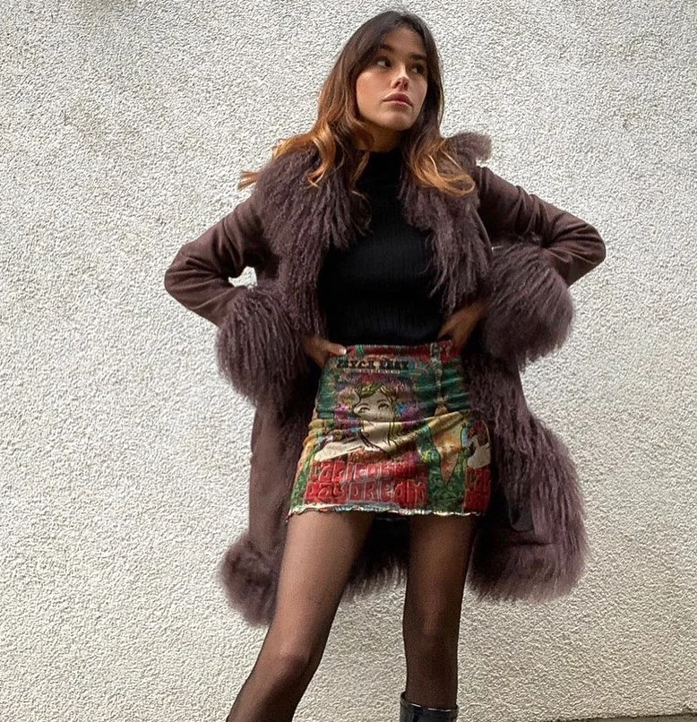 woman standing in front of a wall she is leaning on, looking away from the camera and wearing a fancy brown coat