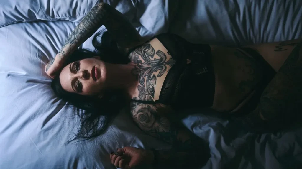 tattooed woman laying in bed at night while getting lit by moonlight, looking seductively into the camera and posing for the night portrait photography