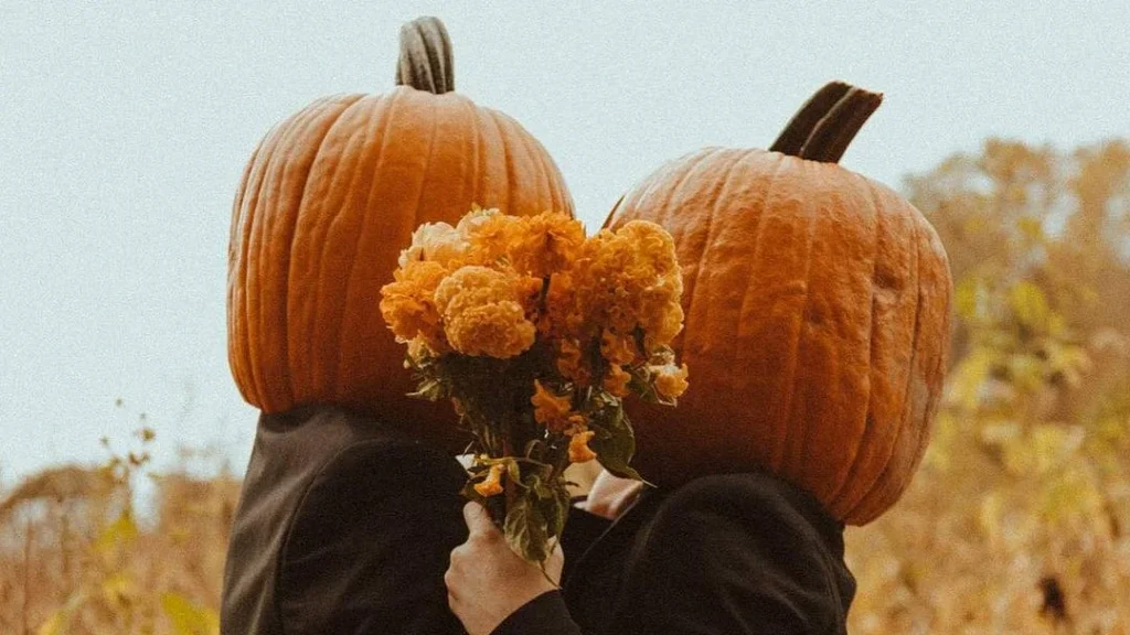 pumpkin heads kissing while flowers are held in the foreground, cute couple doing a pumpkin head couple photoshoot