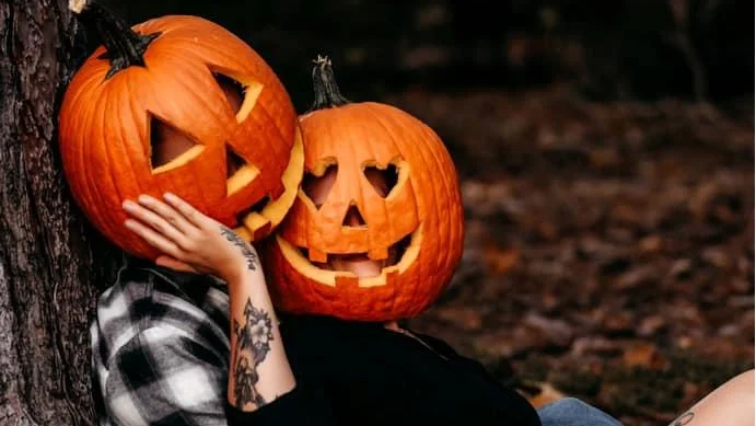 couple sitting next to a tree in a forest wearing pumpkin heads
