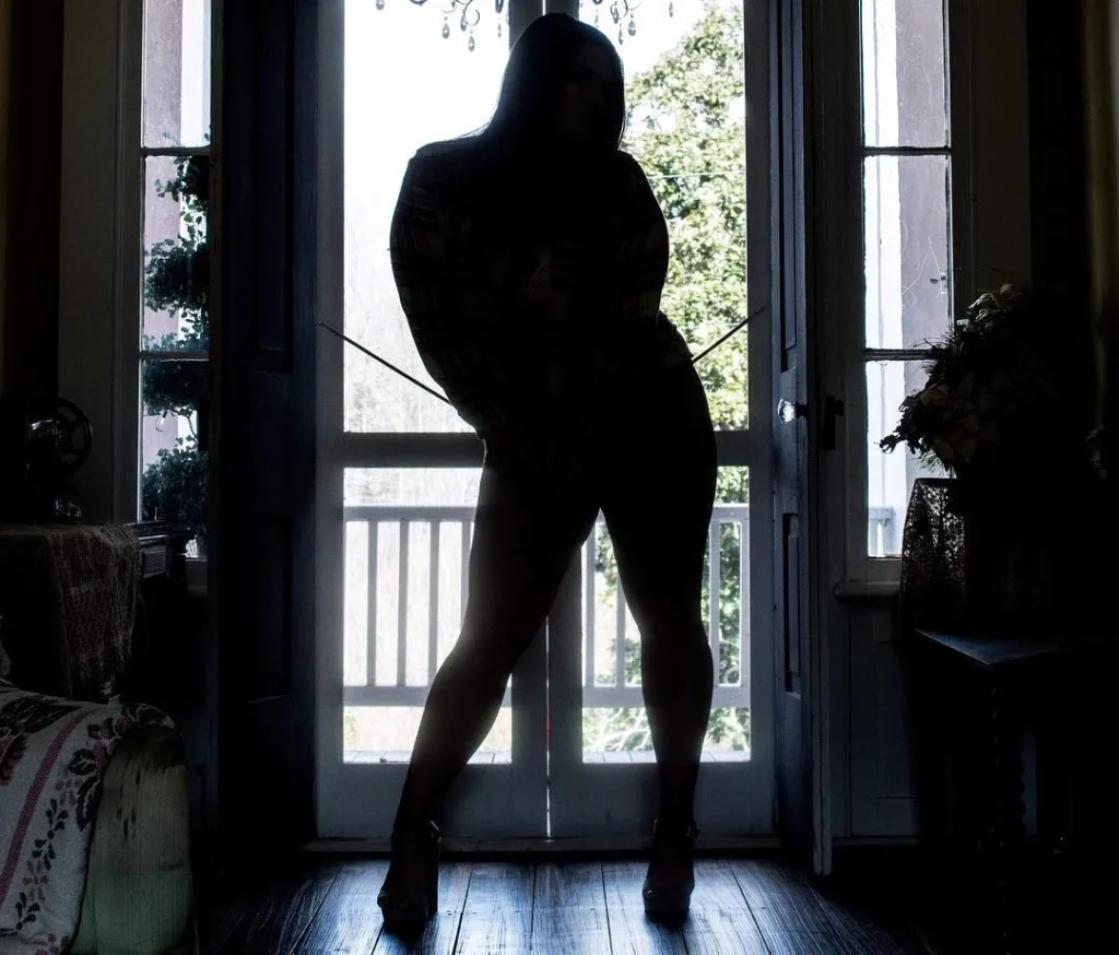 confident woman standing in front of a window with natural light, posing for a boudoir photoshoot in a dark silhouette