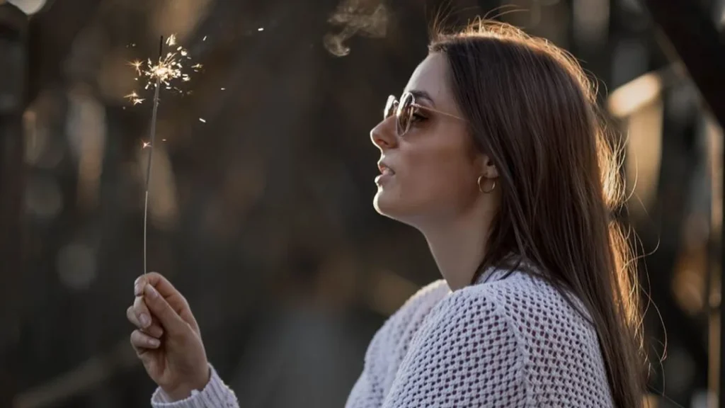 girl holding a sparkler in her hand to pose for a new years photoshoot, wearing white knoted shirt and sunglasses, during the golden hour