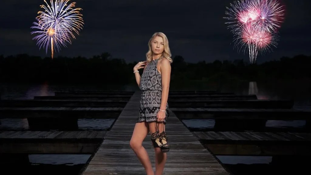 Woman posing on a peer with fireworks in the background, blonde woman, having her shoes in her hands and striking a cute pose