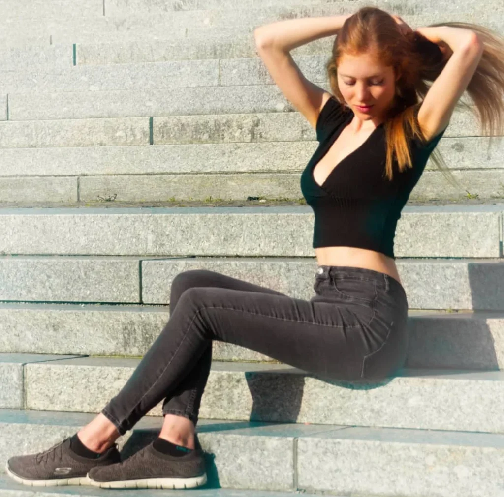 pretty girl with brown hairs making her hair while sitting on stairs wearing a black top and black skinny jeans for a outdoor portrait photoshoot