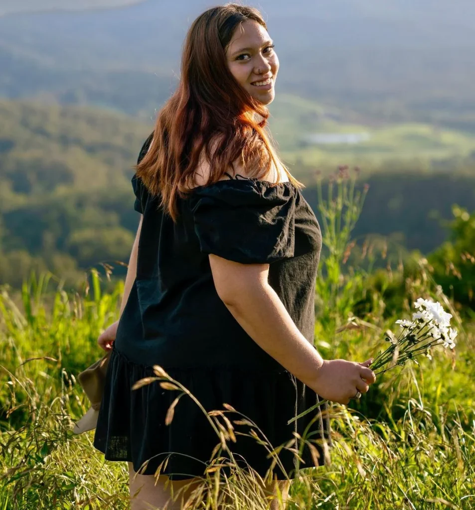 thick girl in black dress holding white flowers in a beautiful green landscape outdoors, smiling and looking at the camera